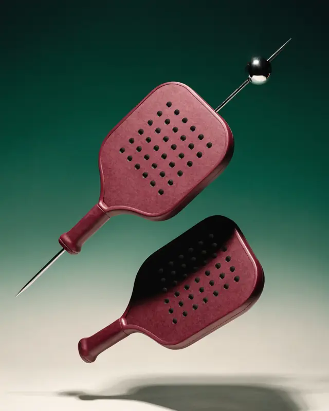 Two burgundy padel paddles threaded on a chrome cocktail pick, floating over a green-to-bone gradient