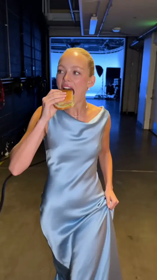 Aurora in a satin dress walking down a studio hallway with a snack, a still from a clip made in AIGE